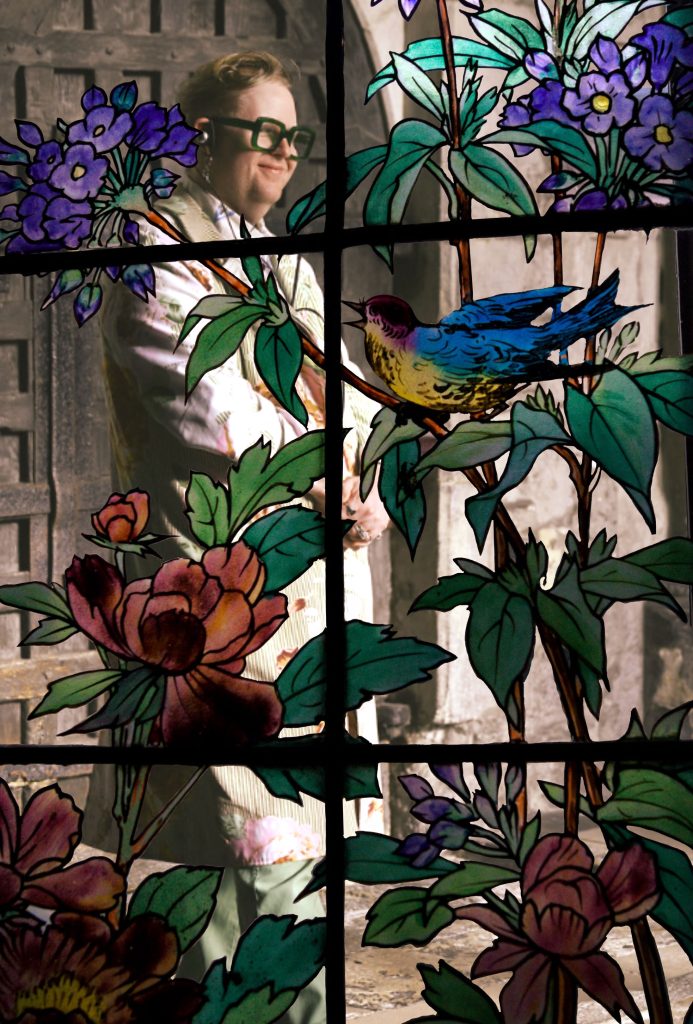 A stained glass window showing a bird in trees and a man in the background.