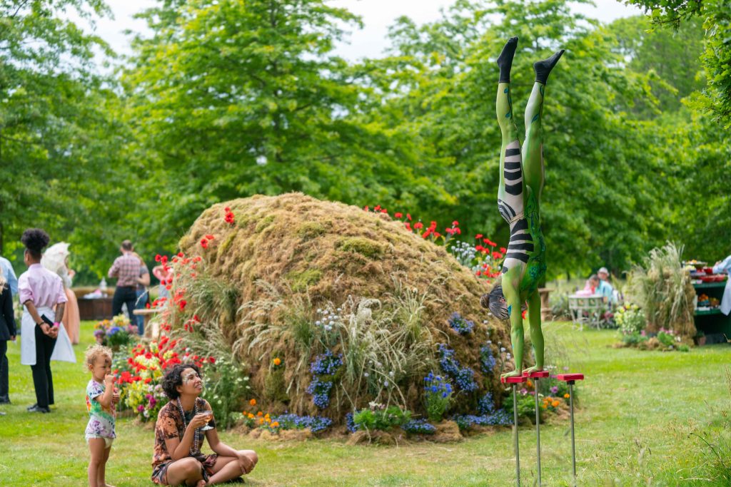 A circus performer balancing on a stand observed by a family in a garden in front of a mound covered in moss and flowers. Luxury party planning.