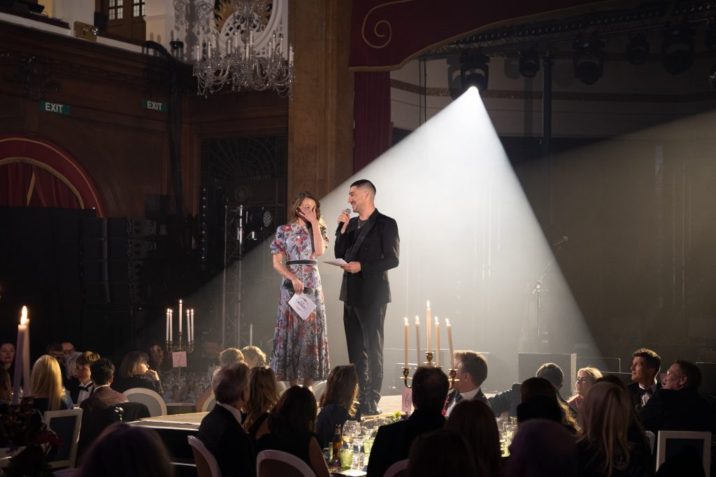 A man and a woman on stage framed by white light with other people sat at tables in an event.