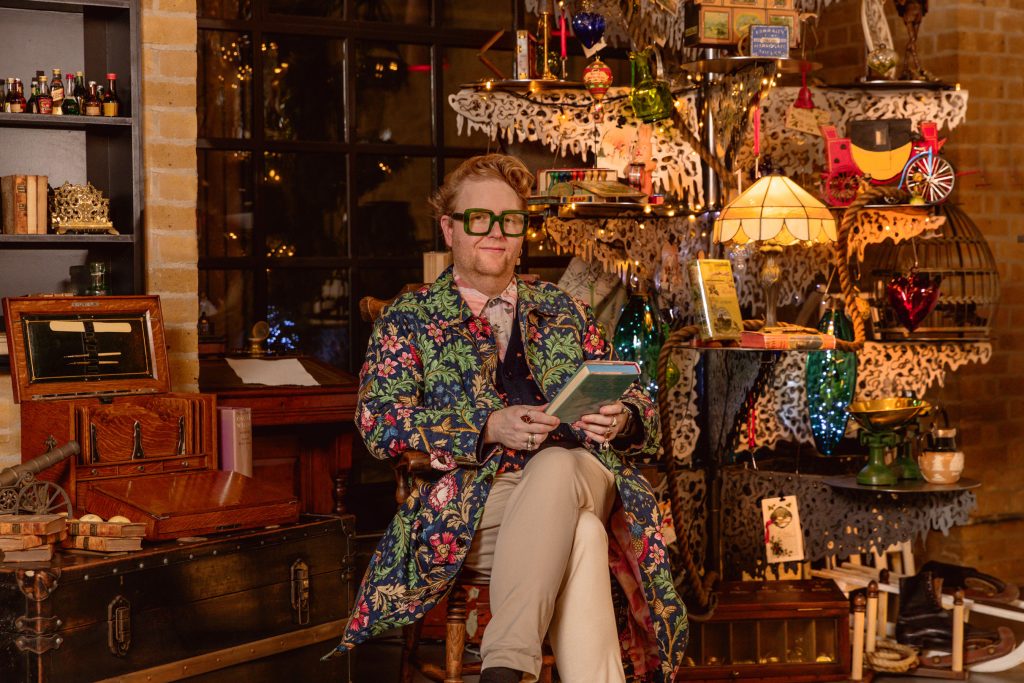 A man wearing a floral print coat reading a book in front of a highly decorated Christmas Tree