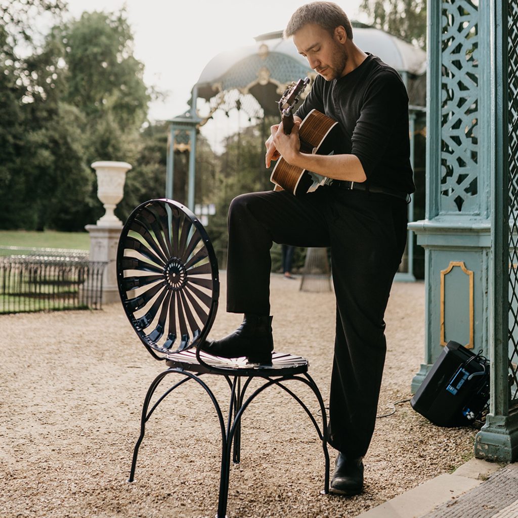 A guitarist wearing black with his knee up on a green metal chair at Waddesdon Manor's Aviary.