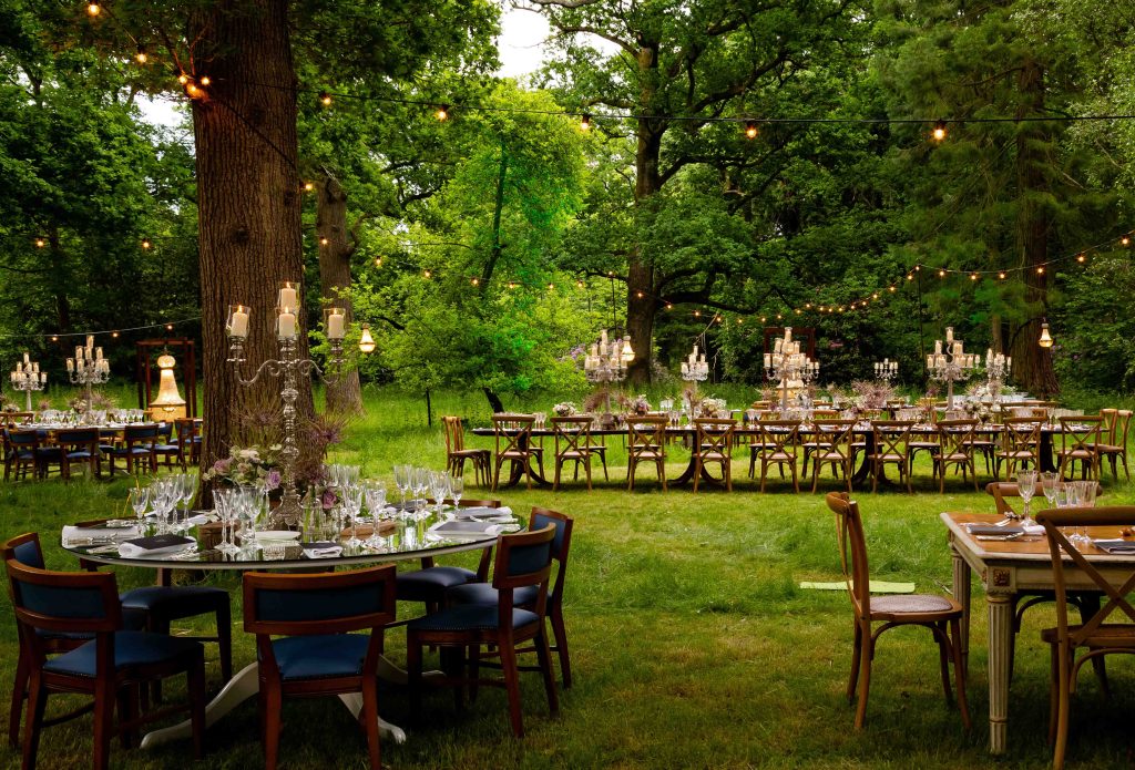 Wooden Dinner tables setup in a woodlands with chandeliers and candles.
