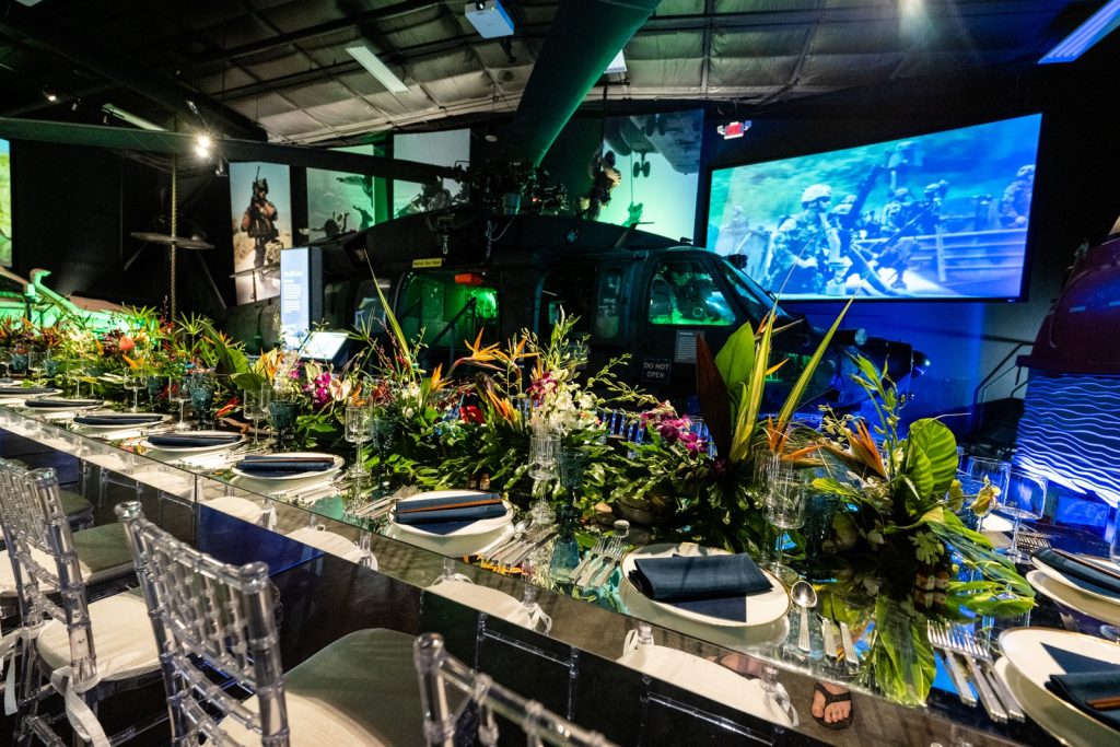 A Black Hawk Helicopter behind a long dining table dressed with exotic flowers and clear acrylic chairs.
