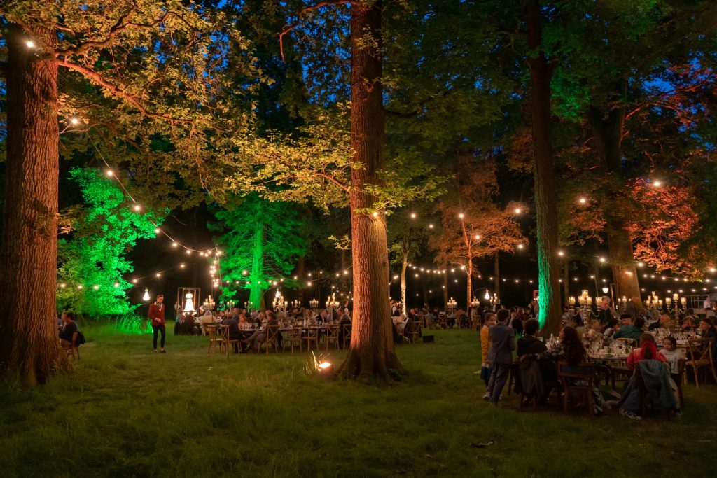 Lots of dining tables positioned in a woodlands dressed with fair lights and green uplighters