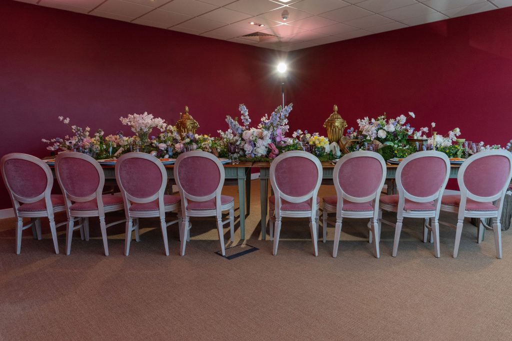 Eight pink velvet chairs set along a table dressed with flowers in a pink painted room. Luxury event planners.