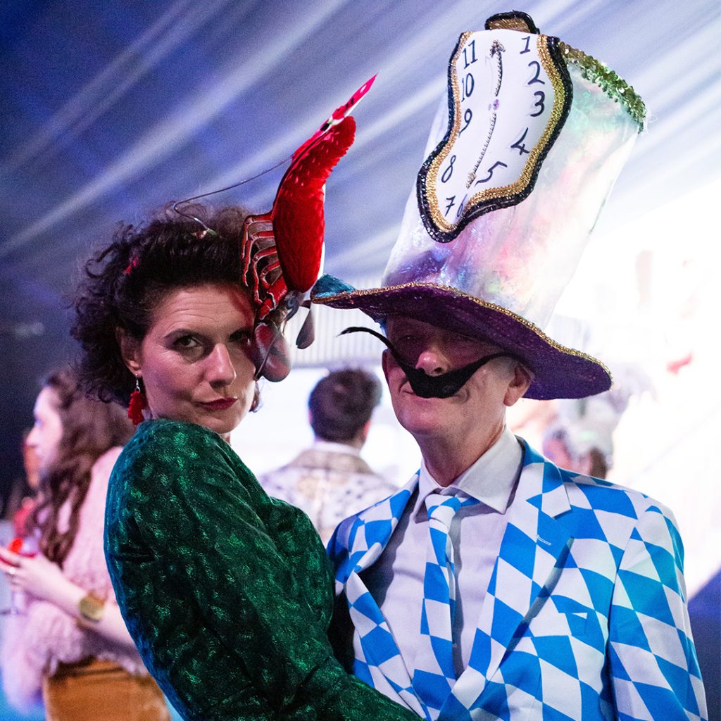 A man and woman wearing fancy hats and colourful, surrealist attire.