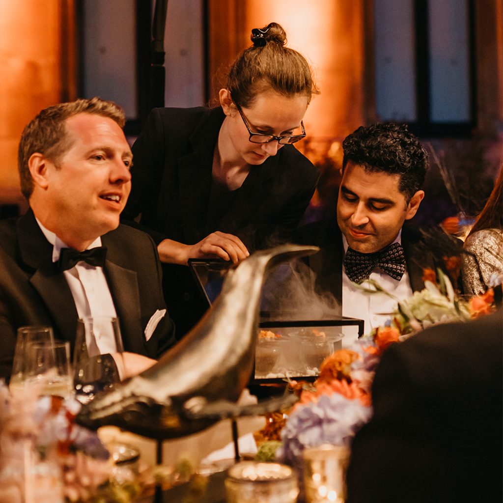 Three people at a dining table ornately dressed with a whale sculpture in the foreground of the photo. A woman holding a box with smoke pouring out of the box. Two guests wearing bowties.