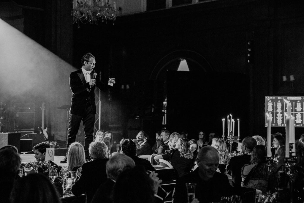 Black and white image of Jude Law on stage in front of people sat at banqueting tables.