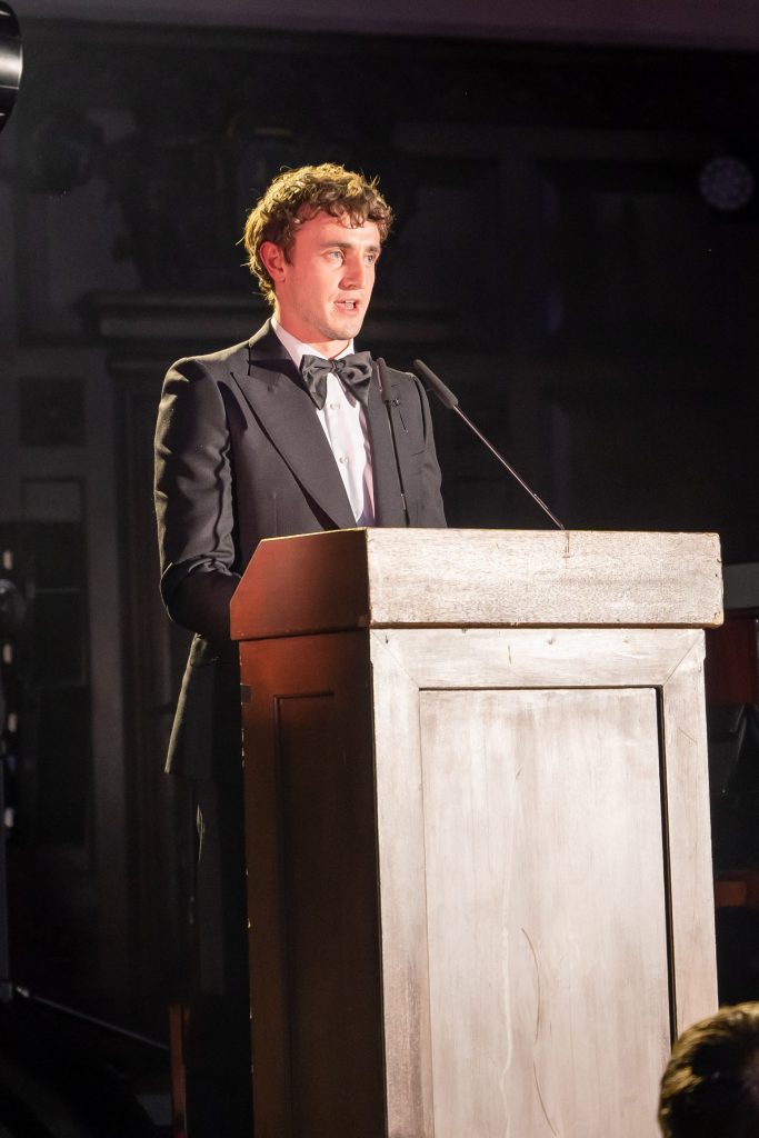 A man in a tuxedo standing behind a wooden lectern presenting on stage.