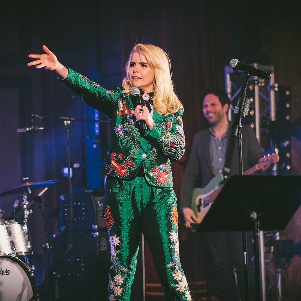 A woman in a green and pink body suit performing on stage with her arm raised.