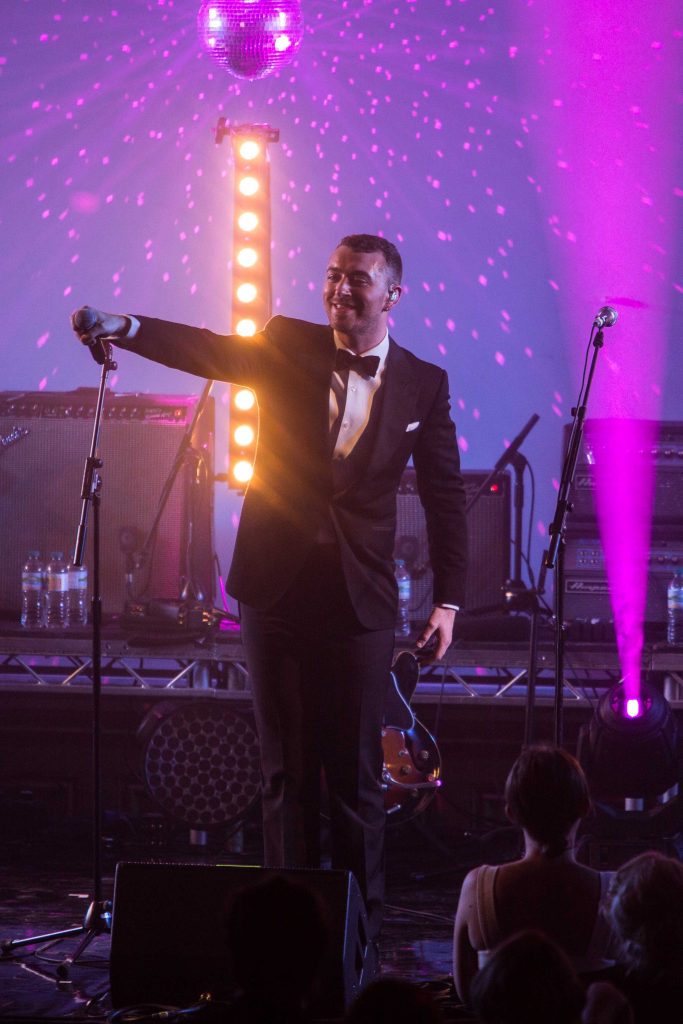 A performer on stage in a tuxedo holding a microphone out to the crowd with purple lighting.