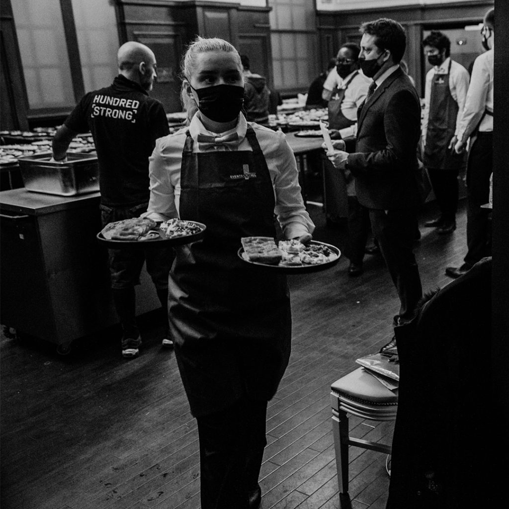 A black and white image of waiters holding plates of food wearing aprons embroidered with the logo 'The Events Meal'