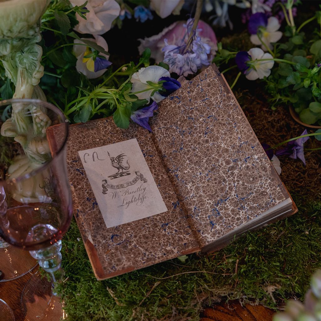 An old book lying flat opened to reveal its inner first page, on a bed of moss and flowers.