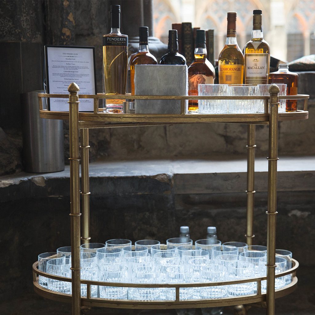 A cocktail station with glassware on the bottom level and spirits and garnishes on the top shelf.