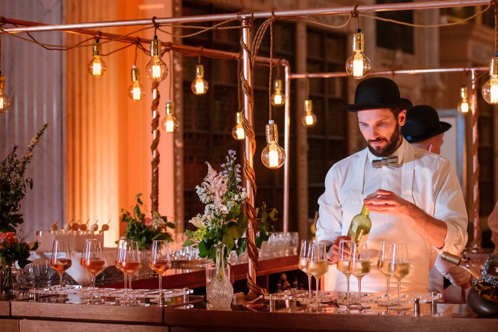 A bar and bartender standing within. Tungsten lighting hanging from copper pipes.