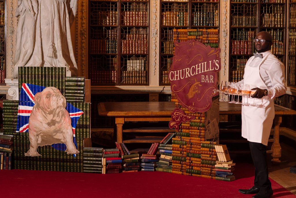 A man holding a tray of drinks in front of two signs, one with a British Bulldog design and another which is a Churchill's Bar sign.