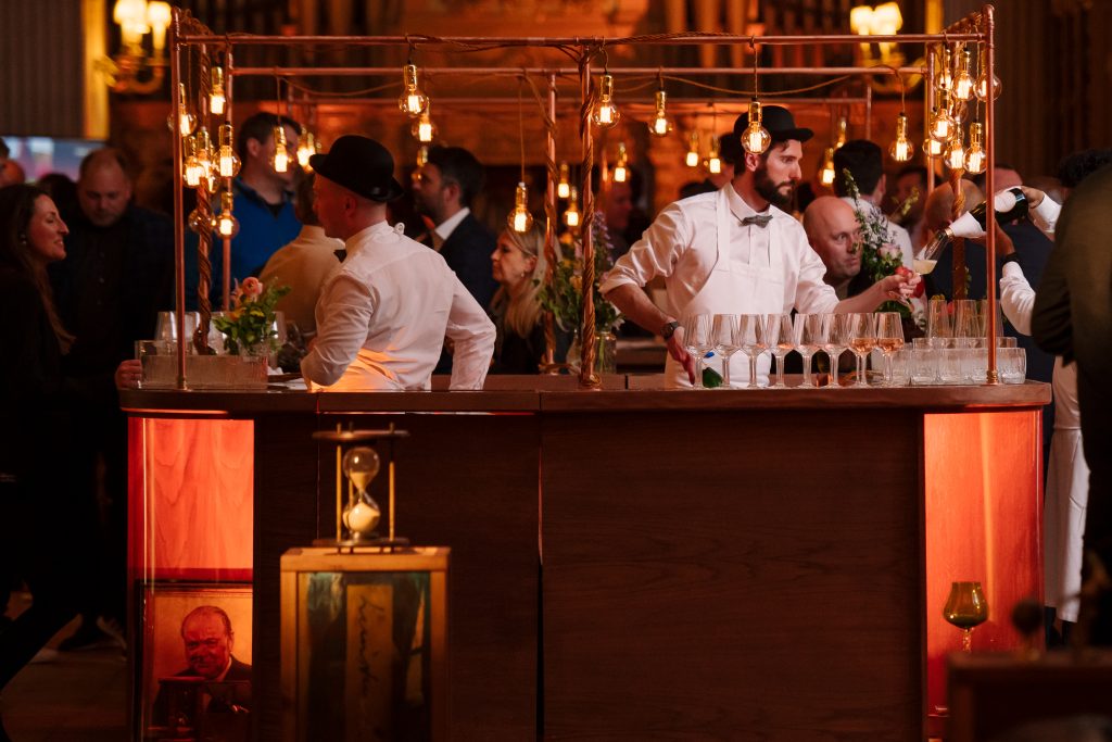 A wooden par with suspended Tungsten lights and bartenders in white aprons and bowler hats.