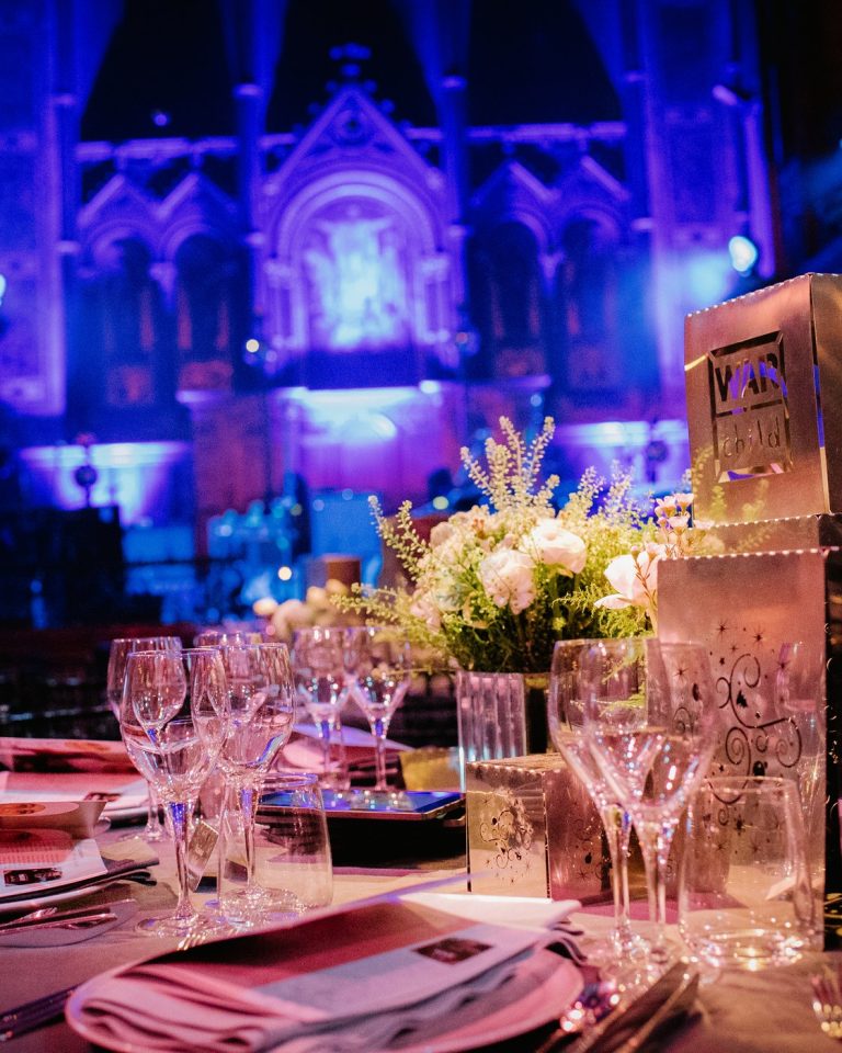 A candlelit table setting with a War Child place marker in front of the blue-lit gothic interior of Christ Church Spitalfields