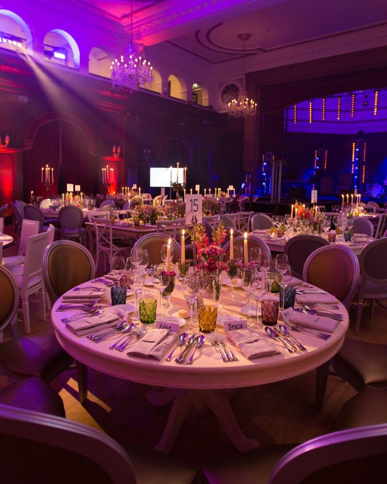 A grand period ballroom with crystal chandeliers and red damask walls, candlelit round tables dressed with florals receding toward a stage