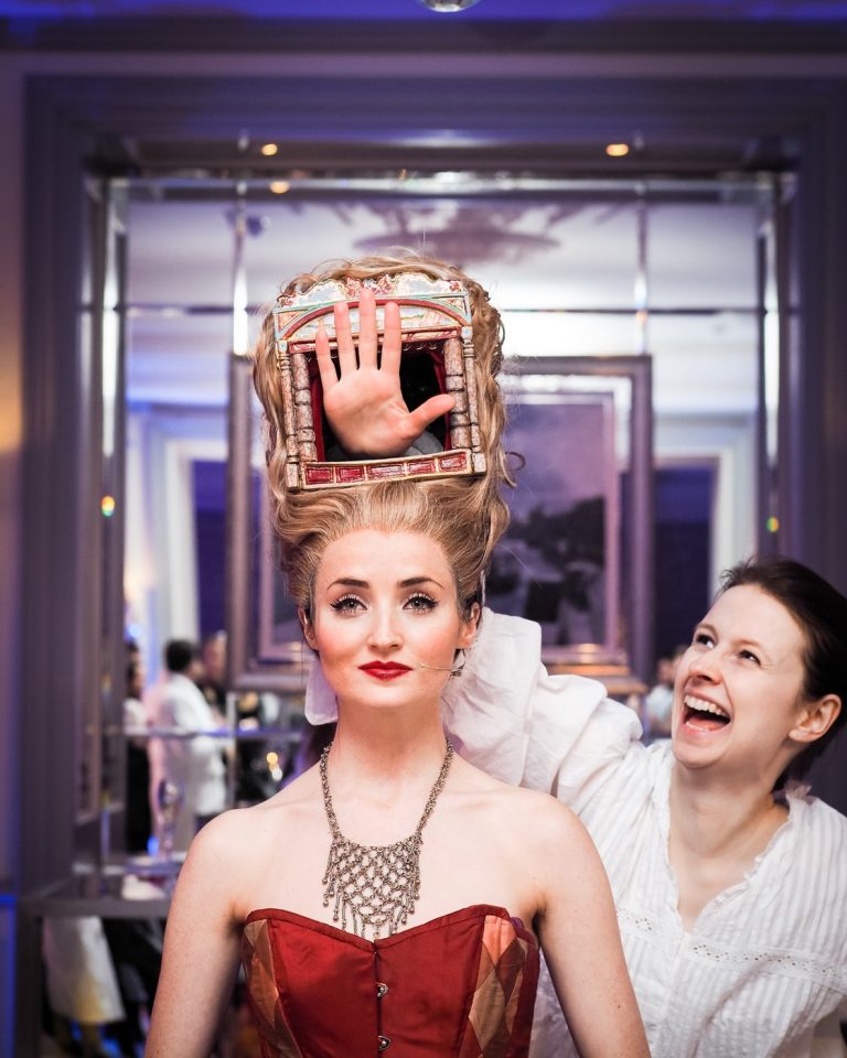 A performer wearing an elaborate headpiece with a working puppet theatre built into it, beside a laughing guest