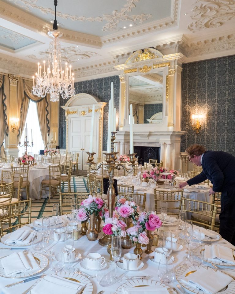 A grand room at Claridge’s with a chandelier, damask walls, a gilt overmantel mirror and gold chiavari chairs
