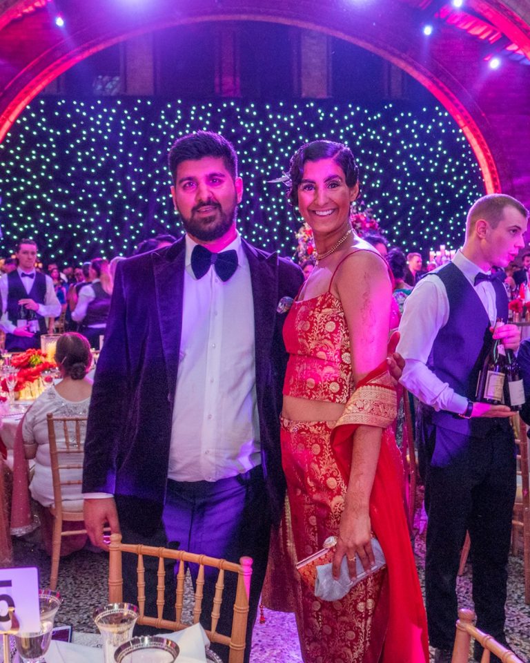 A bride in a red and gold lehenga beside a groom in Western black tie, in front of a fairy-light backdrop