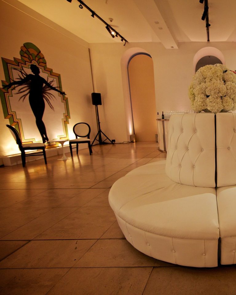 An Art Deco styled lounge with a showgirl silhouette panel, a round white tufted banquette and warm uplighting