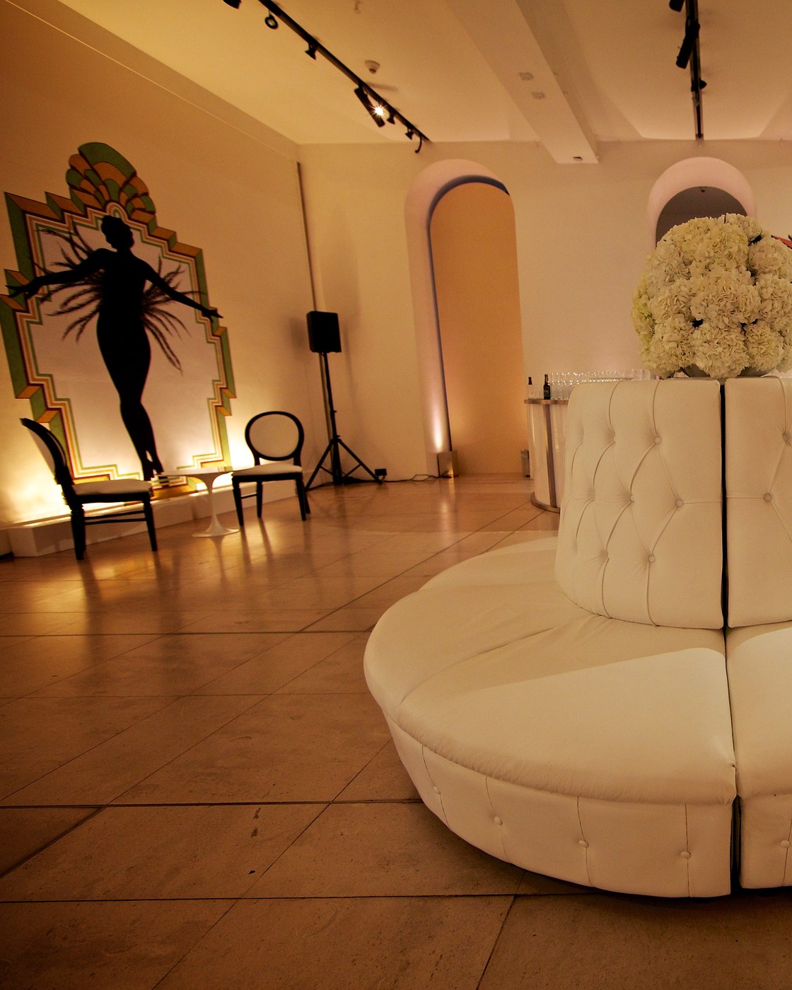 An Art Deco styled lounge with a showgirl silhouette panel, a round white tufted banquette and warm uplighting