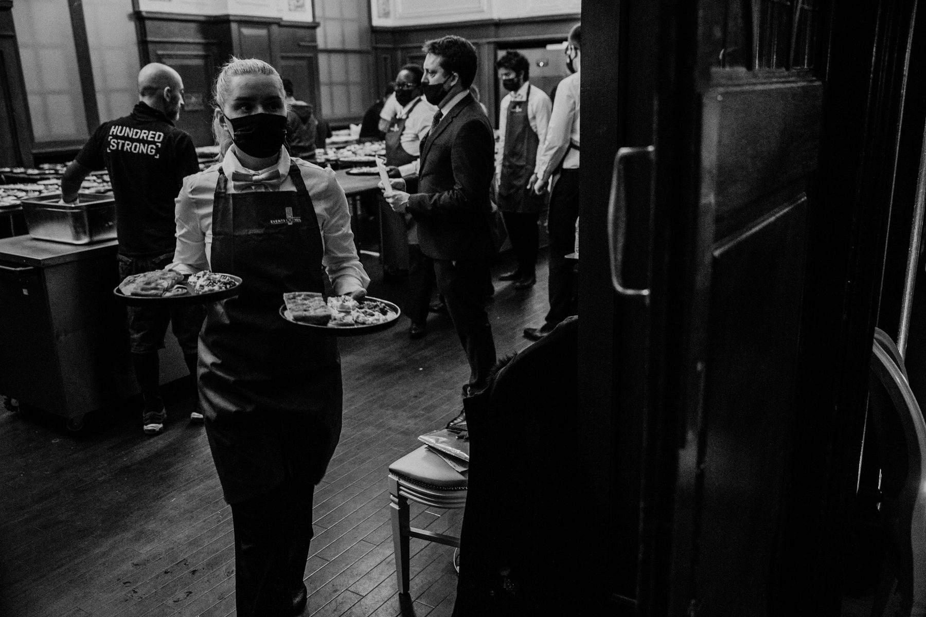 Back of house at the War Child Winter Wassail: two of the crew carrying trays past the kitchen tables, one in a jacket printed Hundred Strong.