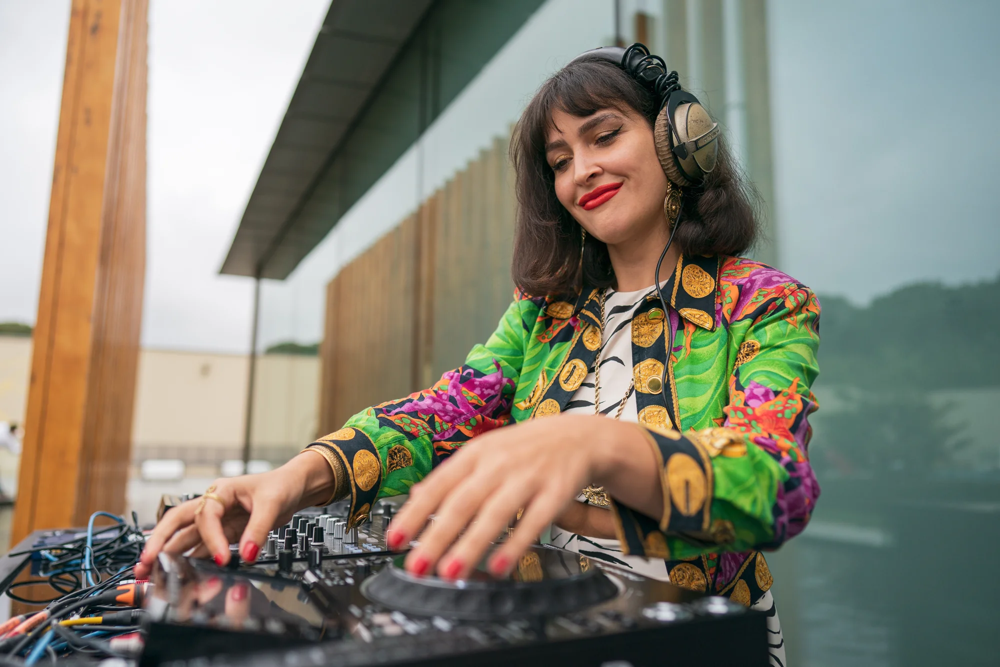The DJ at the decks in headphones and a bright printed jacket, the glass wall of the building behind her.
