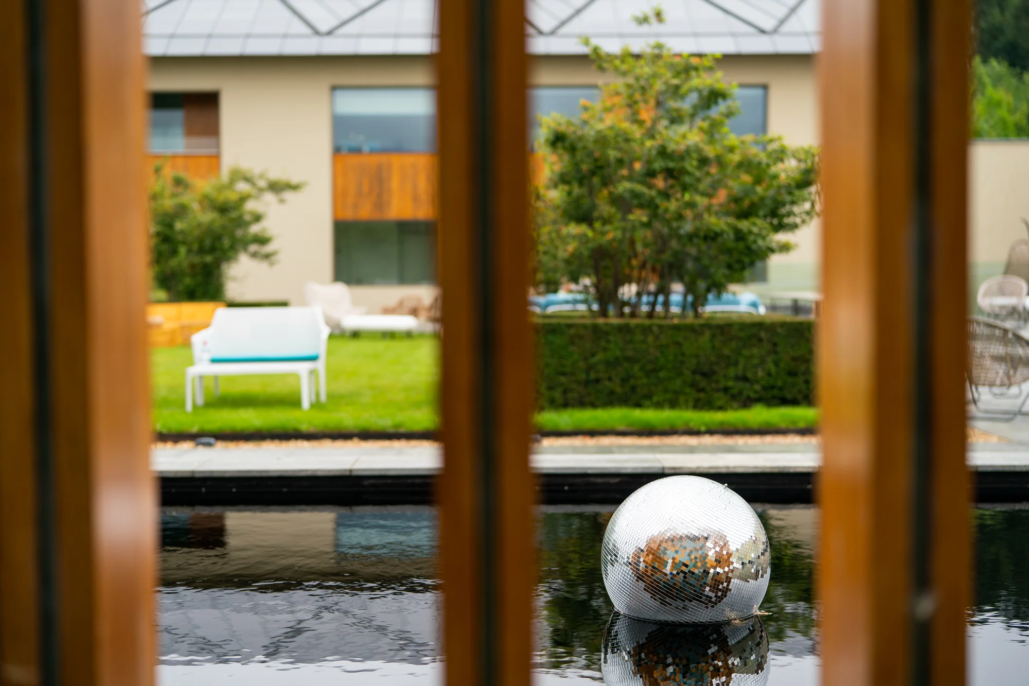 Seen between timber slats, a mirror ball floating on the still water of the pool with the lawns beyond.