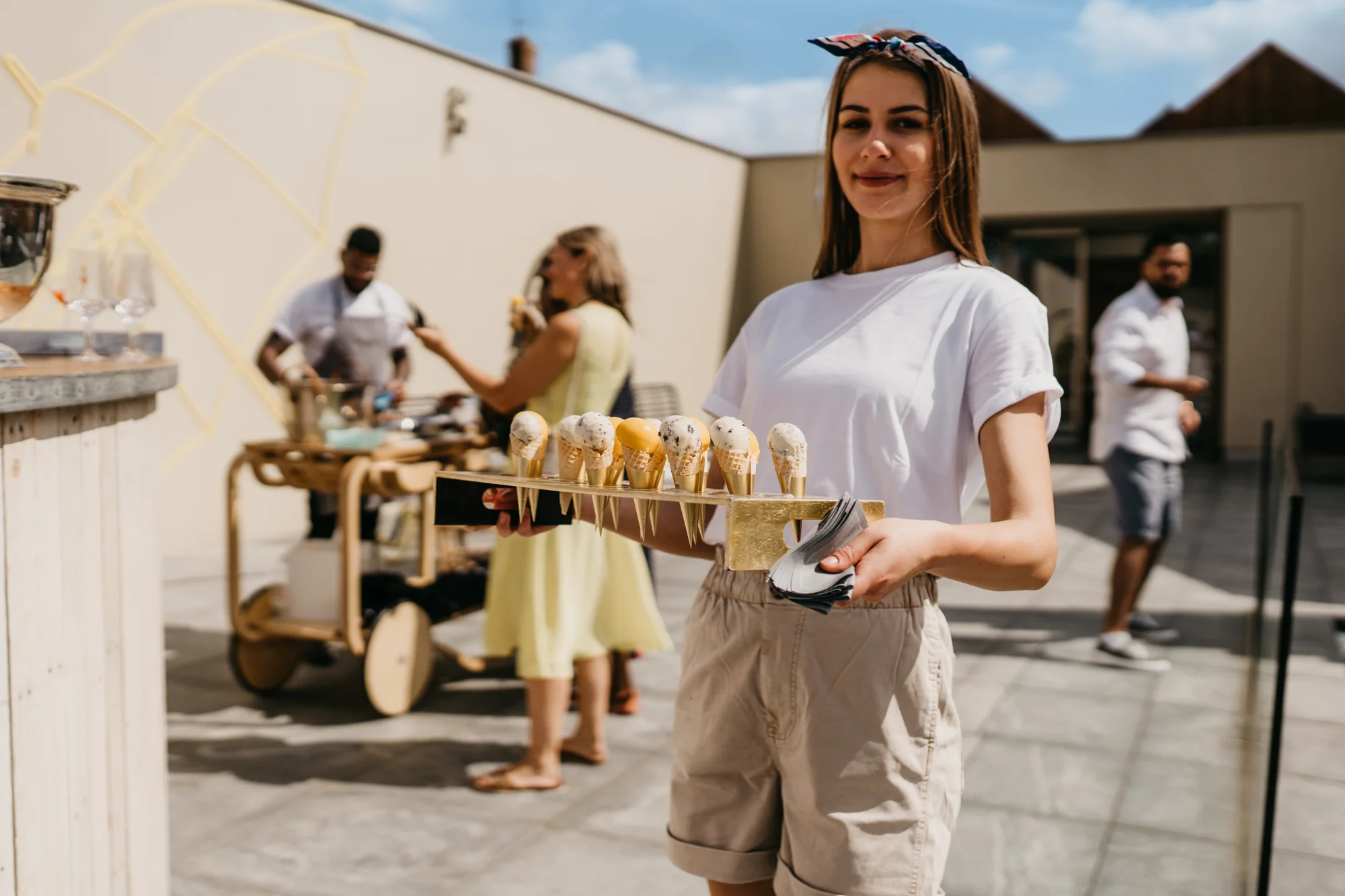 A server carrying an usherette tray of drinks across the terrace, a drinks trolley behind her.