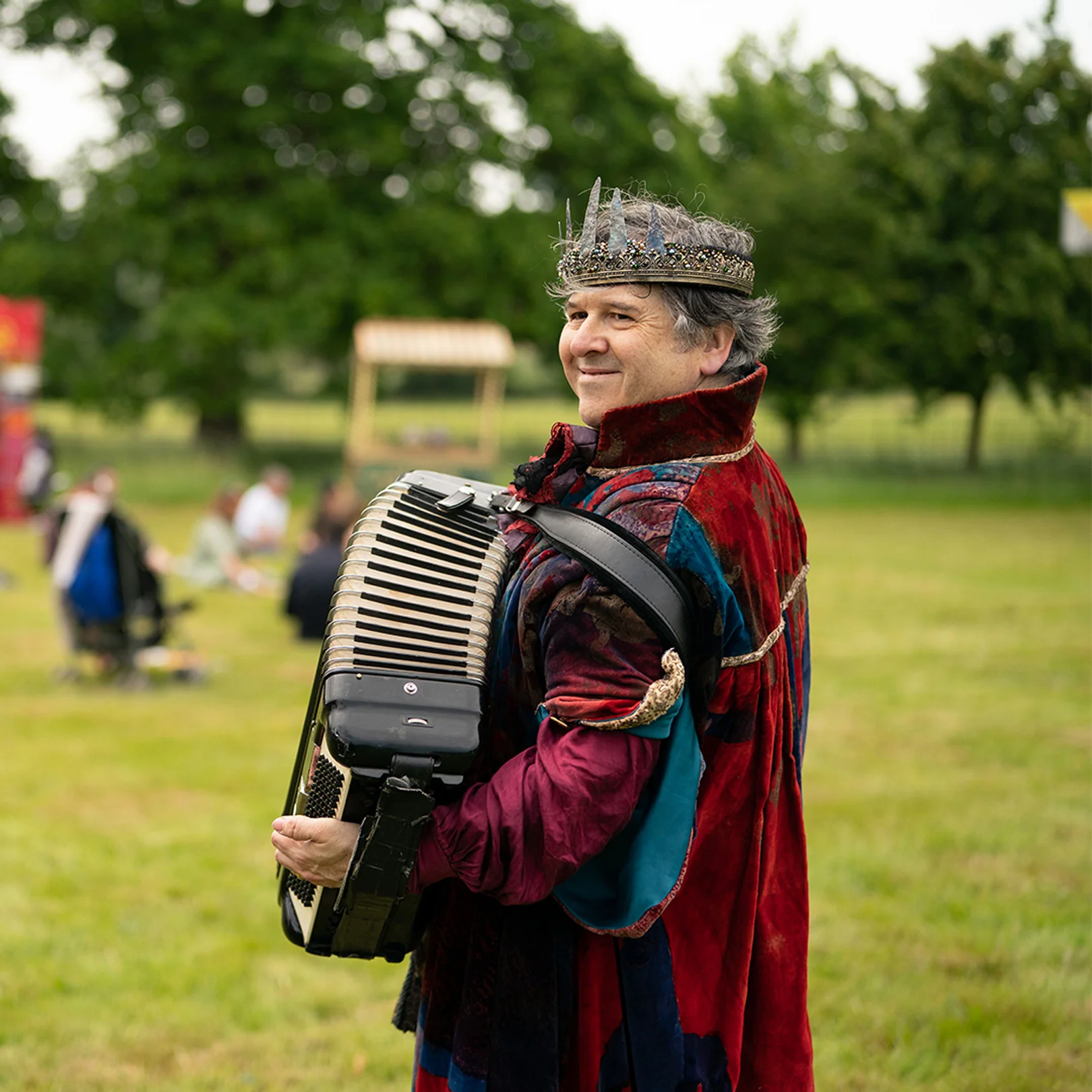 An accordion player in a crown and a red velvet robe walking the field, guests behind him.