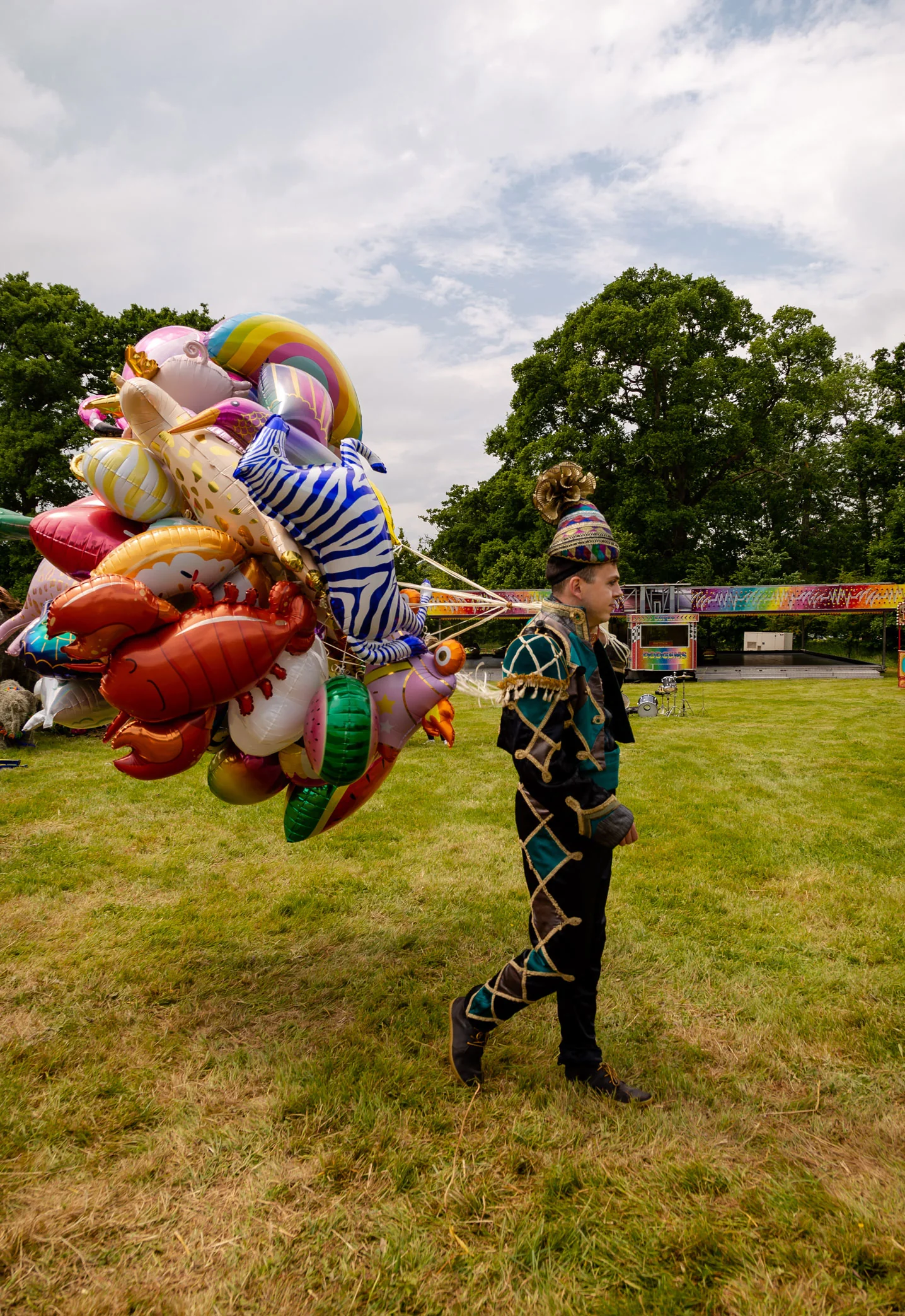 A balloon artist in a harlequin suit carrying a great bunch of balloon animals across the grass.