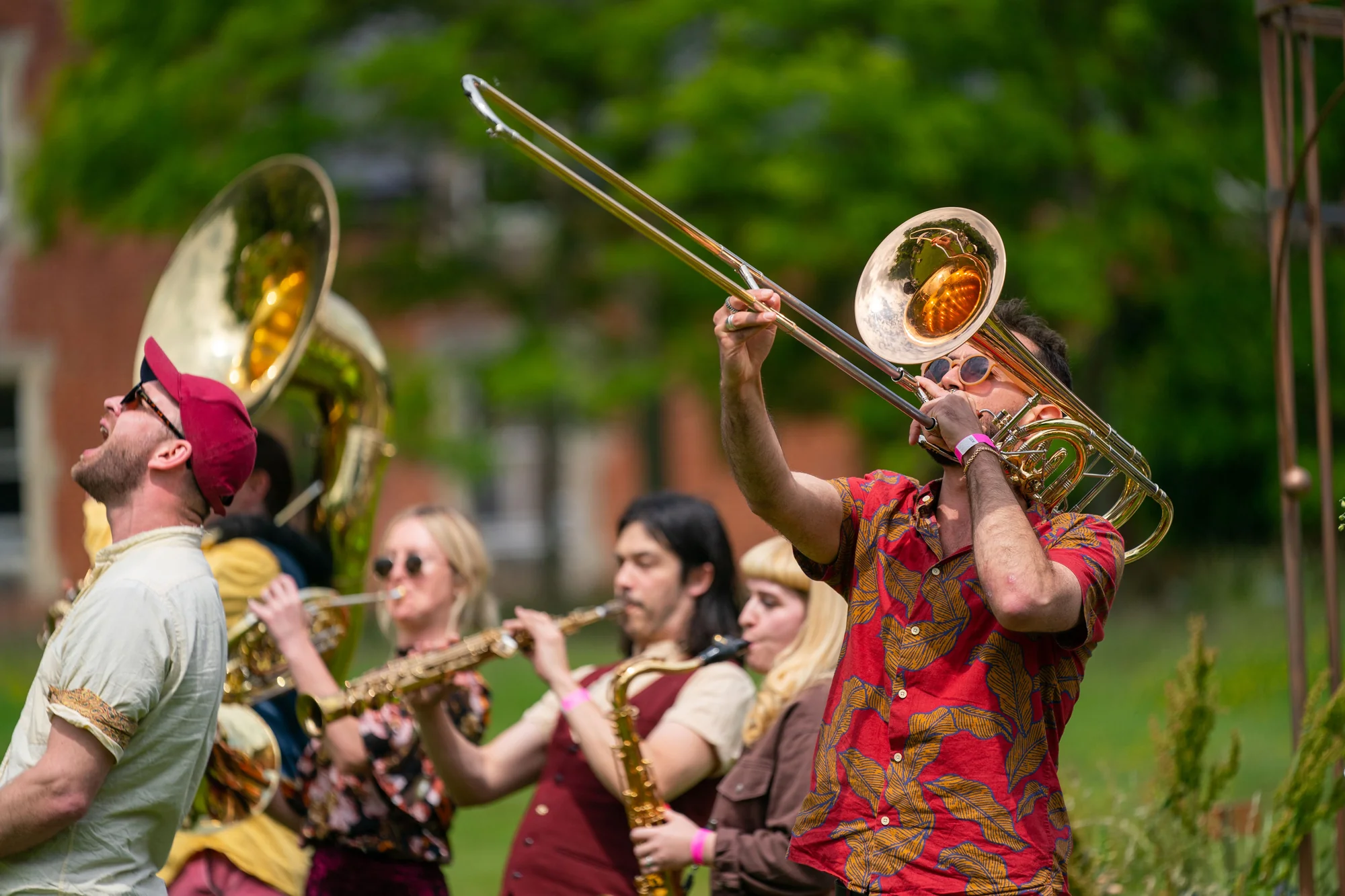 A brass band playing out on the grass, sousaphone and trombone raised, guests gathered around them.