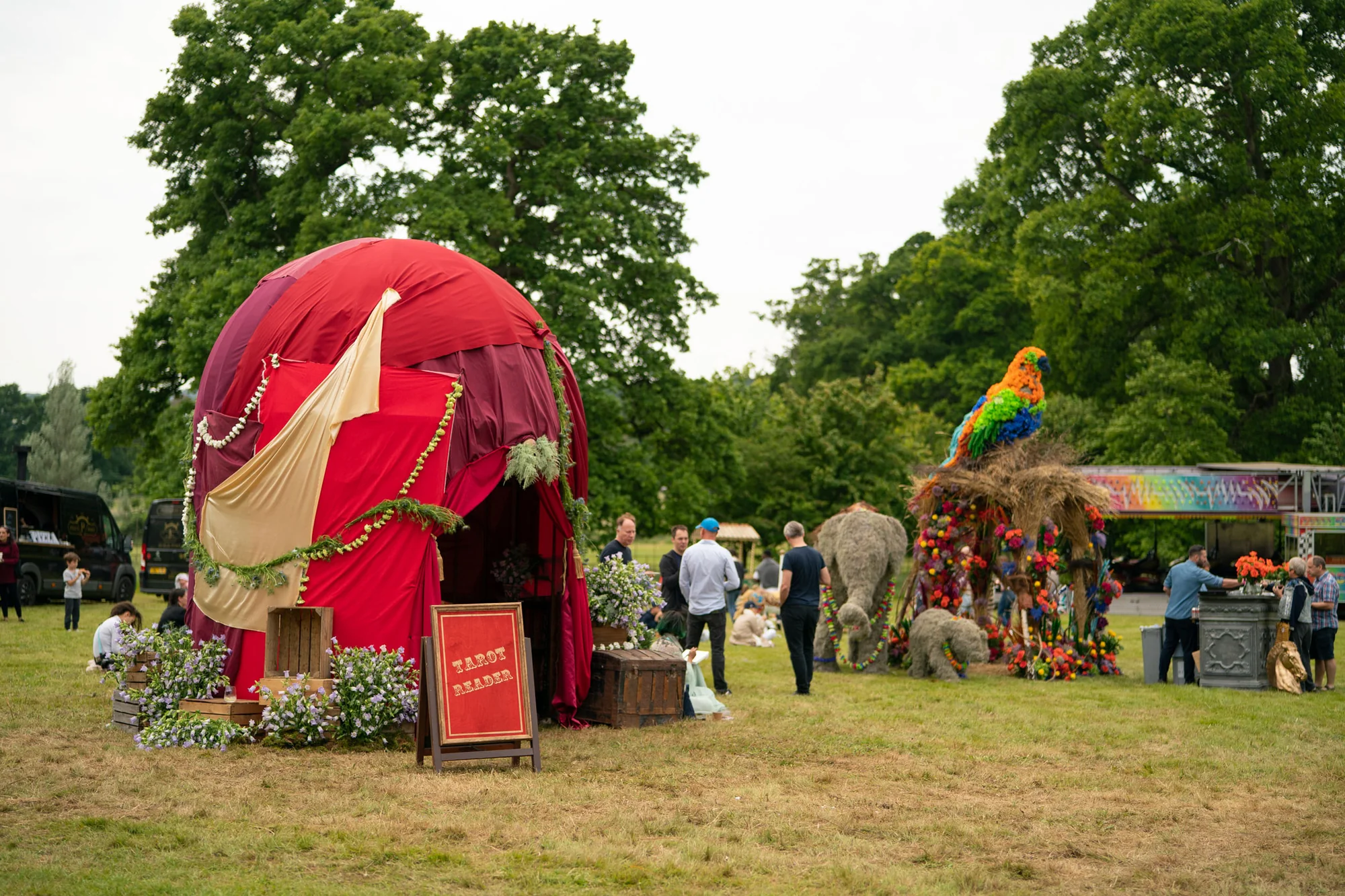 A red and cream circus tent pitched on the grass with the funfair rides and the straw elephant behind it.
