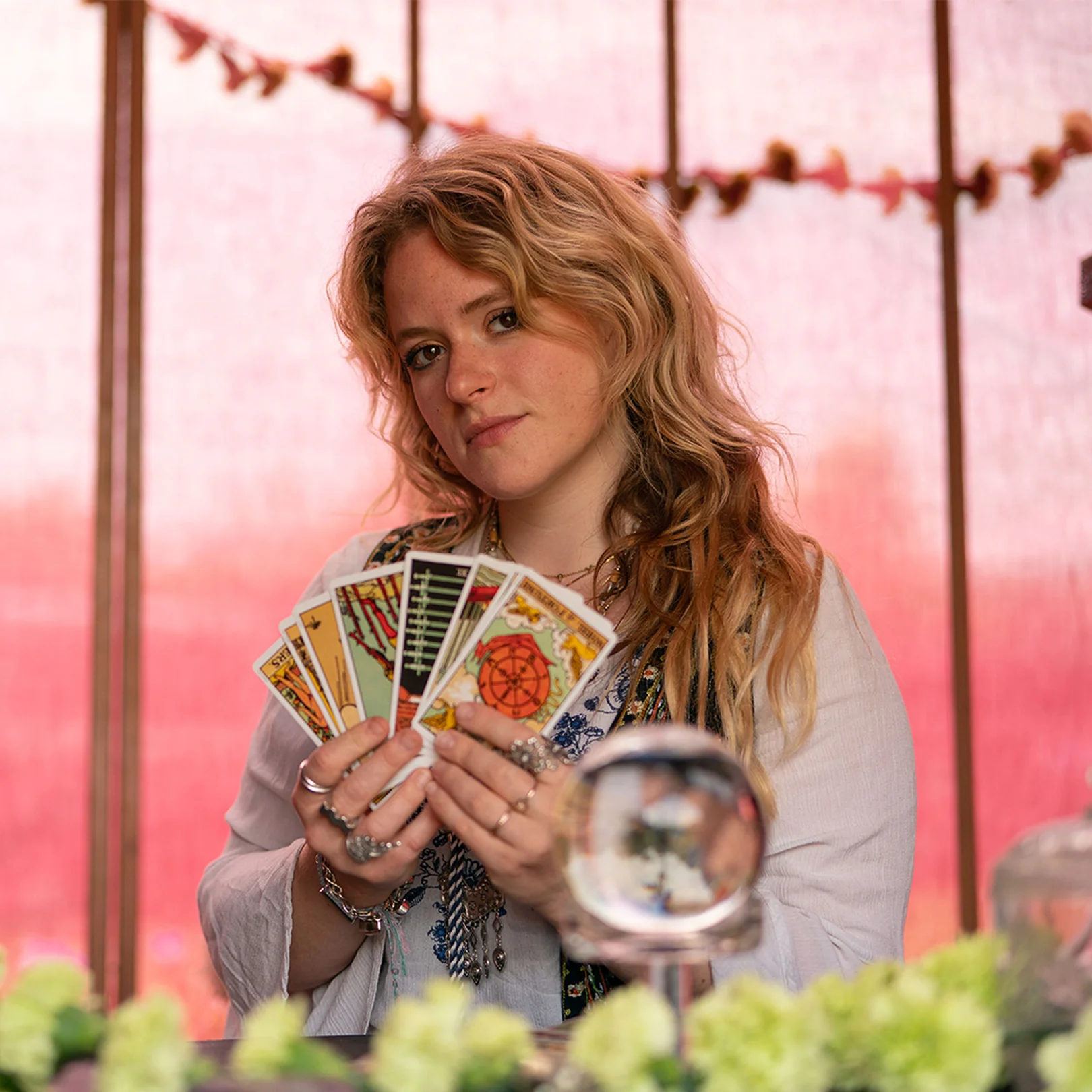 A tarot reader holding a fan of cards and a crystal ball in front of a pink backdrop.