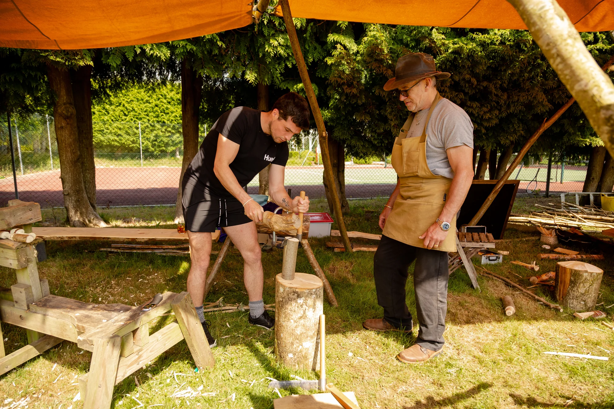 Two guests whittling at trestles under an open canopy, shavings on the ground around them.