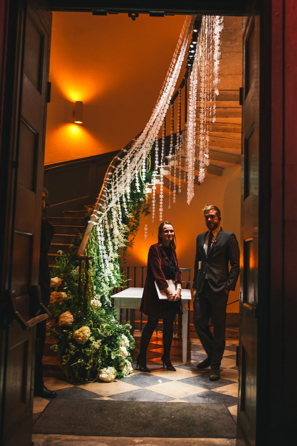 Two guests in the doorway beneath a lit garland running up the staircase.