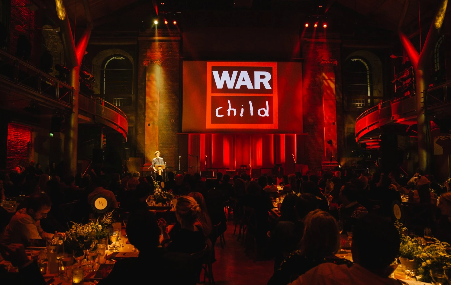 Jerwood Hall full of guests, with the War Child screen lit red between the brick piers.