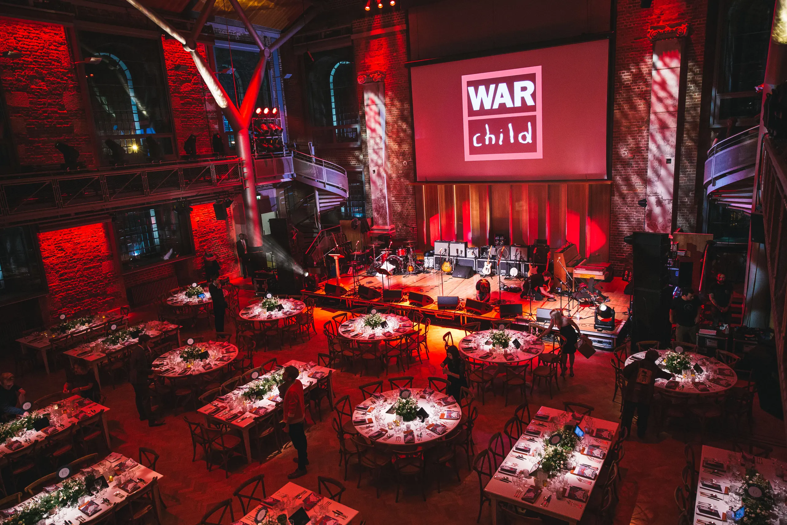 Jerwood Hall at LSO St Luke’s lit red, the whole room seen from the balcony with long and round tables, the stage and the iron galleries on both sides.