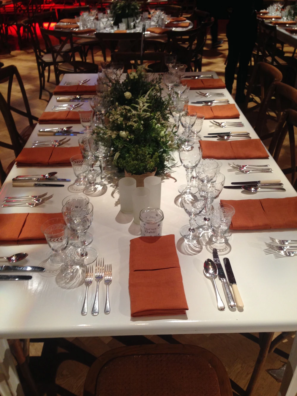 A dressed table with orange napkins, cut glass and a low arrangement of white flowers and herbs.