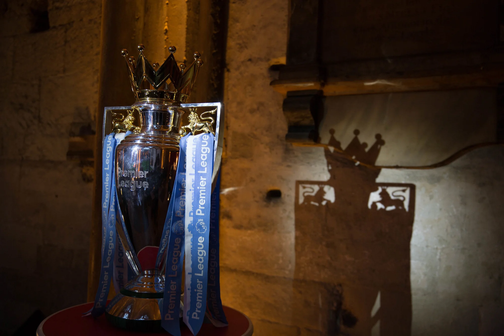 The Premier League trophy lit on its plinth against bare stone, its shadow thrown across the wall behind it.