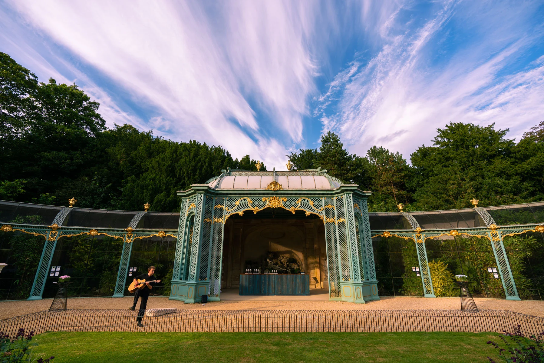 The pale green ironwork of the Aviary at Waddesdon Manor under a wide evening sky, guests gathered at its centre.