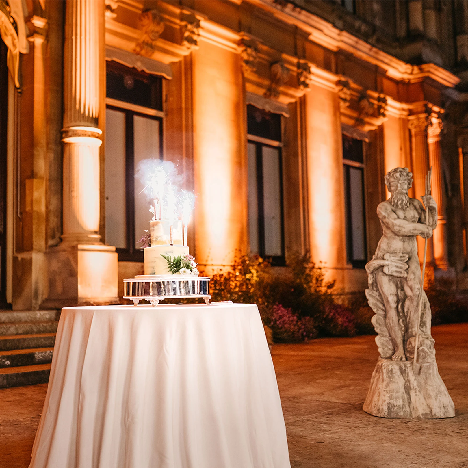 A birthday cake on a draped table beside a stone statue, a sparkler lit on top and the facade uplit behind.