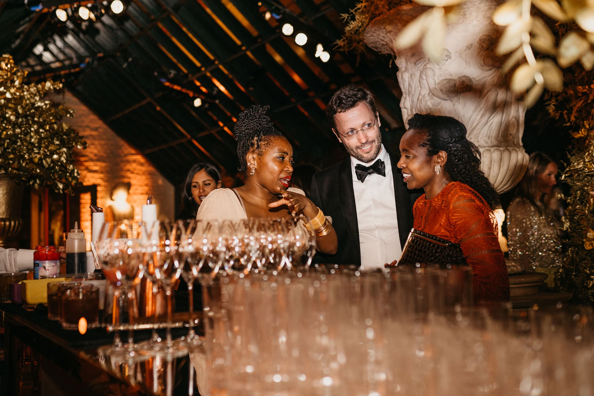 Rows of champagne coupes on the bar with guests talking behind them.