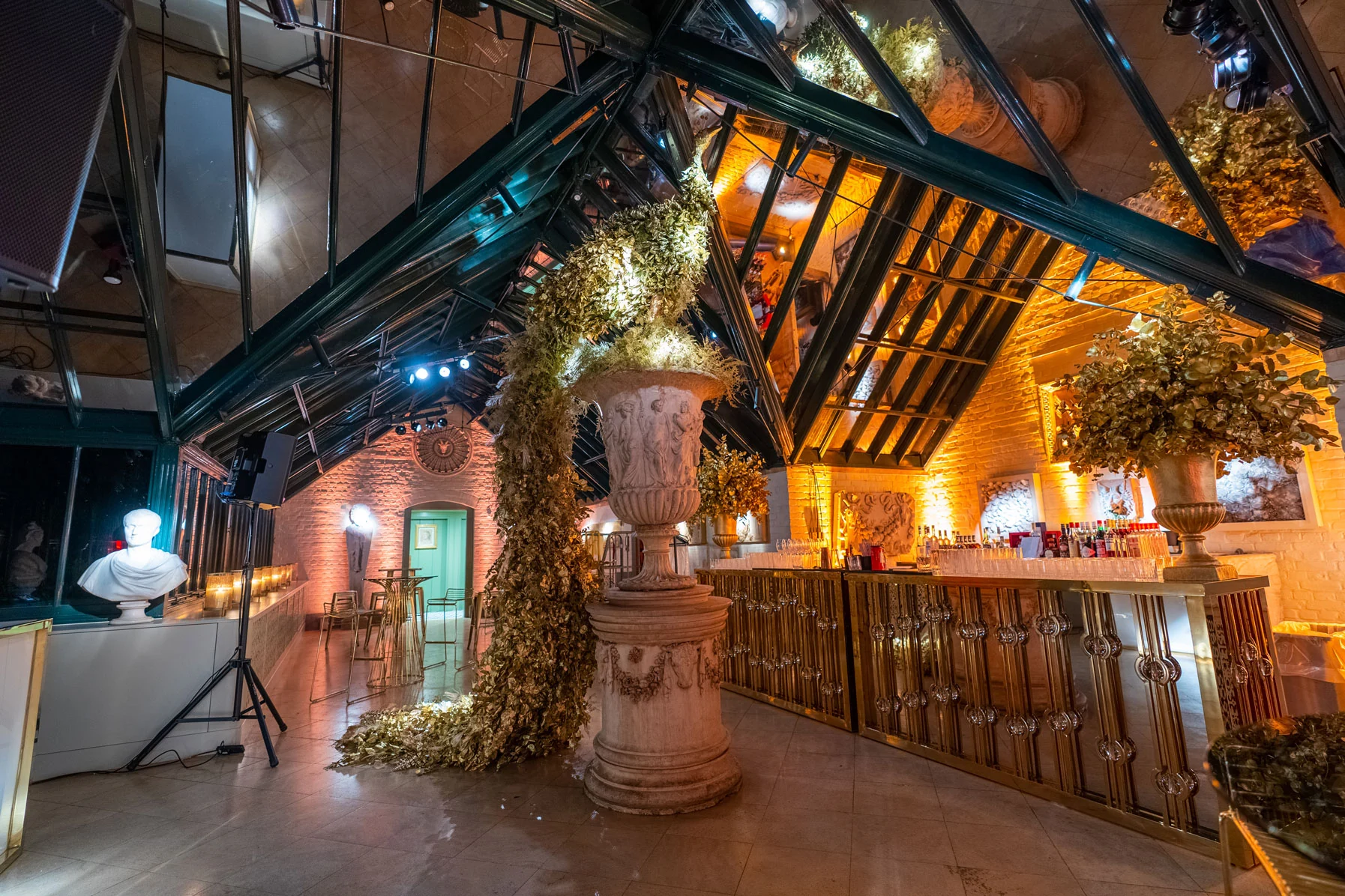 A bar built under a high timber and glass roof, greenery and busts along it, lit green and gold.