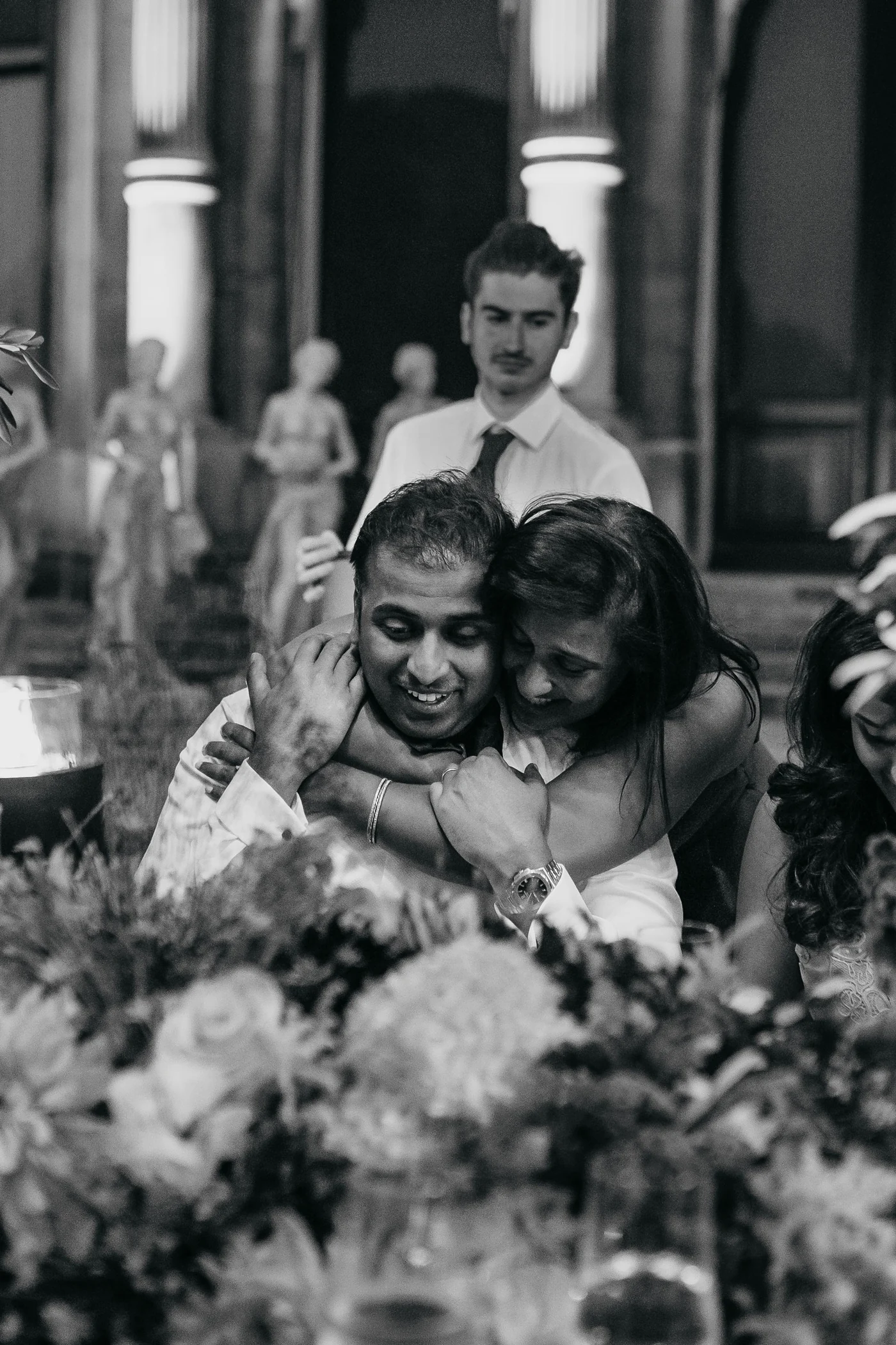 In black and white, two guests holding each other and laughing at the table.