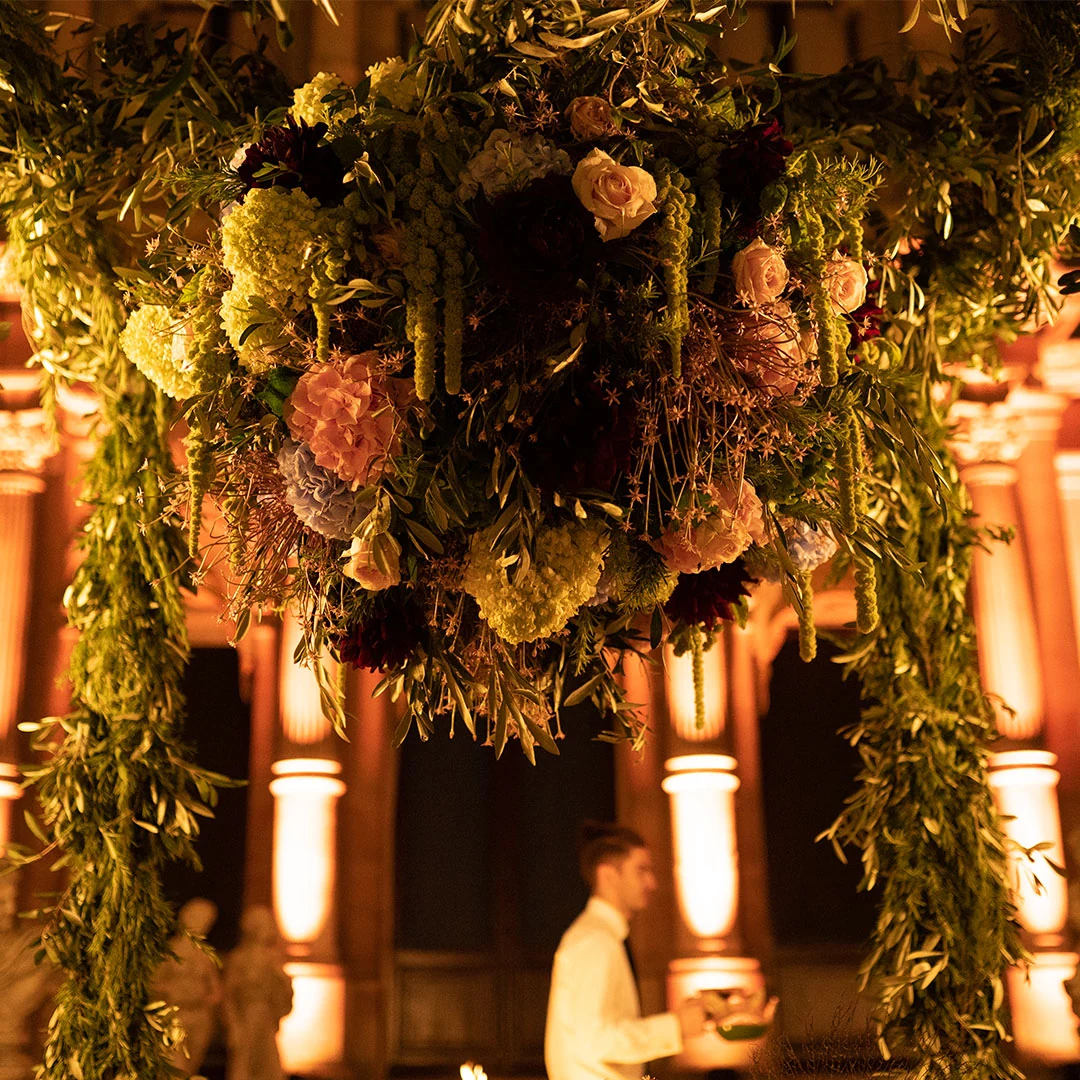 A canopy of roses, dried flowers and trailing foliage suspended above the table, the lit facade behind it.