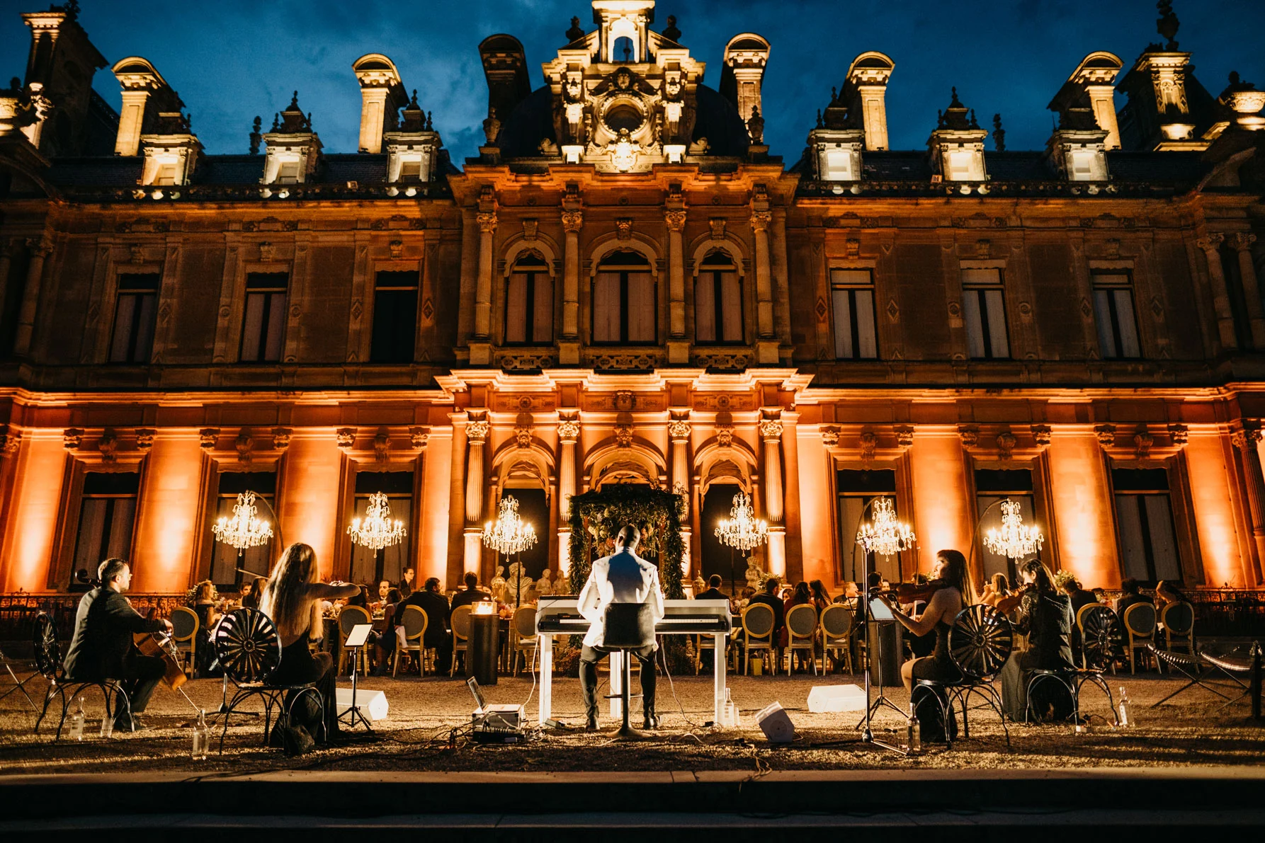 A long table laid down the Parterre at Waddesdon Manor at dusk, the facade uplit amber behind it and a band playing at the far end.